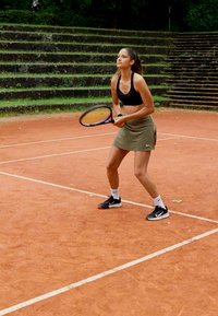 Jugadora de tenis en una cancha de tierra batida, vistiendo un top corto negro y una falda verde, sosteniendo una raqueta, con zapatos deportivos negros y calcetines blancos.