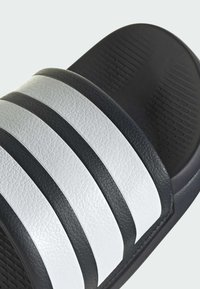 Čierne šľapky so tromi bielymi textúrovanými pruhmi, s hladkým zvrškom a drážkovanou podrážkou pre lepšiu priľnavosť.