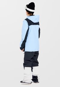 Sneeuwjack in lichtblauw met een zwarte bovenkant, gecombineerd met zwart-witte broek. Beschikt over een capuchon en ritszakken.