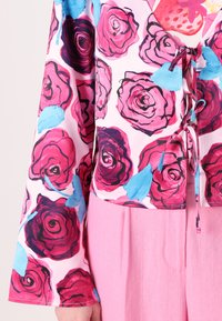 Floral blouse featuring hand-painted pink roses with blue accents on a light pink background, tied at the front with a bow. Bell sleeves.