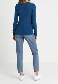 Blauwe kabelgebreide trui met een aansluitende pasvorm, gecombineerd met lichtblauwe denim jeans en witte sneakers. Gebreide textuur en geribde manchetten zichtbaar.