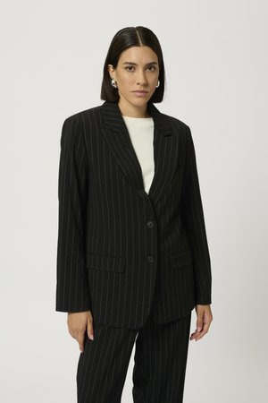 Blazer nero a righe con revers, due bottoni e tasche frontali. Il tessuto ha una texture liscia, abbinato a un top crema sottostante.
