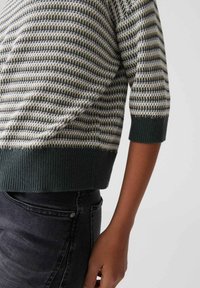 Braccio e vita di una persona che indossa un maglione a righe verdi e bianche con maniche a tre quarti e jeans grigio scuro.