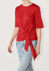 Top rosso a maniche corte con dettaglio annodato sul davanti, realizzato in tessuto morbido. Abbinato a jeans chiari strappati, con una vestibilità ampia.