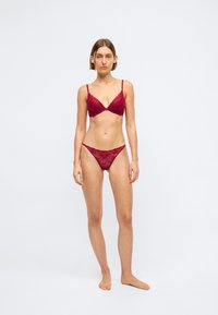 Femme debout pieds nus portant un ensemble assorti en dentelle rouge, comprenant un soutien-gorge et une culotte, sur un fond blanc uni.