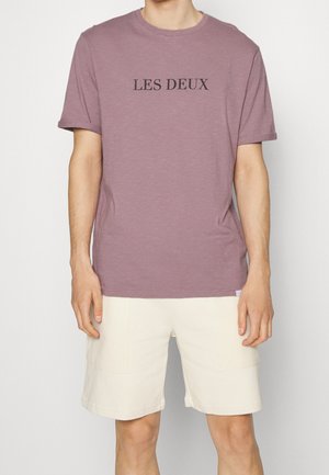 T-shirt en coton violet avec l'inscription "LES DEUX" en noir sur la poitrine, associé à un short beige clair avec poches latérales.