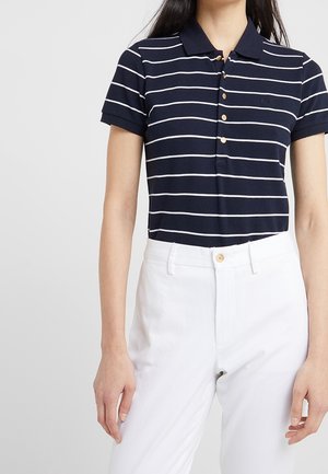 Vrouw draagt een marineblauw met wit gestreept polohemd met korte mouwen, ingestopt in een witte broek met hoge taille, tegen een effen achtergrond.