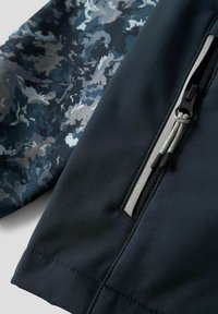 Veste imperméable en tissu bleu marine foncé avec une manche à motif camouflage dans des nuances de bleu et de gris. Poche zippée avec des accents gris.