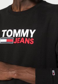 Sudadera negra con el texto "TOMMY JEANS" en blanco y rojo. Presenta un cuello redondo y puños con detalles del logo en rojo, blanco y azul. Textura suave.