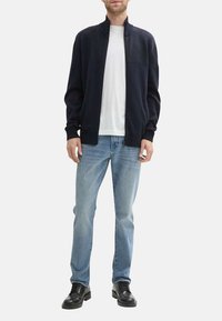 Ciemnoniebieski sweter z dzianiny z teksturowanym górnym częścią, dopasowanymi mankietami i stójką, noszony na białej koszulce i jasnoniebieskich dżinsach.