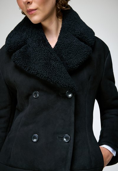 Femme portant un manteau noir croisé avec un grand col texturé et les mains dans les poches, légèrement tournée vers la gauche.