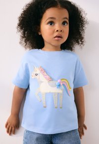 T-shirt en coton bleu clair arborant un motif de licorne avec des détails brodés, une crinière rose et des pompons colorés. Manches courtes, coupe décontractée.