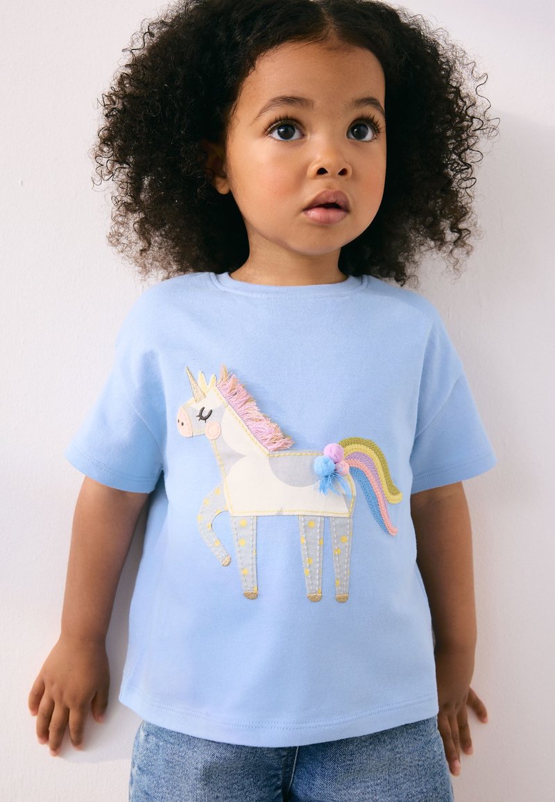T-shirt en coton bleu clair arborant un motif de licorne avec des détails brodés, une crinière rose et des pompons colorés. Manches courtes, coupe décontractée.