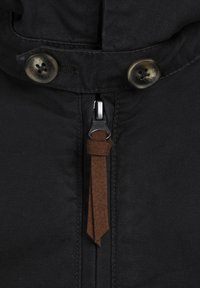 Black fabric jacket close-up featuring a metal zipper with a brown faux leather pull tab and two round, brass buttons.
