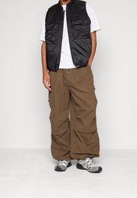 Gilet in nylon nero con numerose tasche indossato sopra una maglietta bianca, abbinato a pantaloni cargo beige larghi e scarpe da ginnastica grigie.