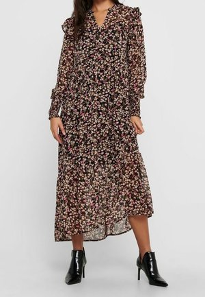 Frau trägt ein langes, transparentes schwarzes Kleid mit floralem Muster, Rüschendetails und langen Ärmeln, kombiniert mit schwarzen Stiefeletten mit hohen Absätzen.