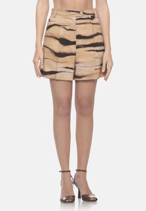 Frau trägt beige High-Waist-Shorts mit schwarzen Tigerstreifen und braune offene High-Heel-Sandalen, steht vor einfachem Hintergrund.