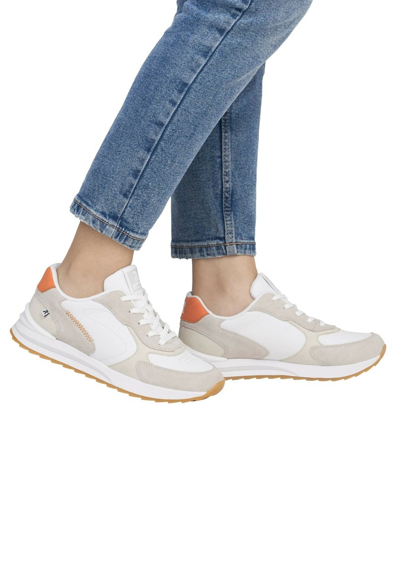 Weiße und beige Sneaker mit Wildlederakzenten, ausgestattet mit einem orangefarbenen Fersentab, Schnürdesign und Gummisohle, getragen mit hochgekrempelten Jeans.
