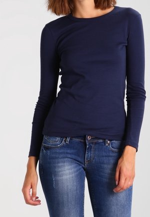 Vrouw die een getailleerd marineblauw overhemd met lange mouwen en blauw verwassen spijkerbroek draagt, staat met één hand ontspannen en de andere iets gebogen.