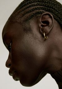 Pendiente de aro dorado en una oreja perforada, llevado por una persona con cabello trenzado. Vista cercana de la textura suave de la piel, ángulo de perfil.