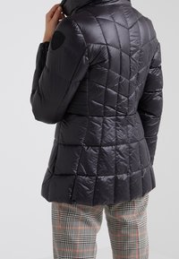 Femme ajustant le col d'une veste matelassée noire avec des coutures géométriques, portée avec un pantalon à carreaux noir et blanc avec des rayures rouges.