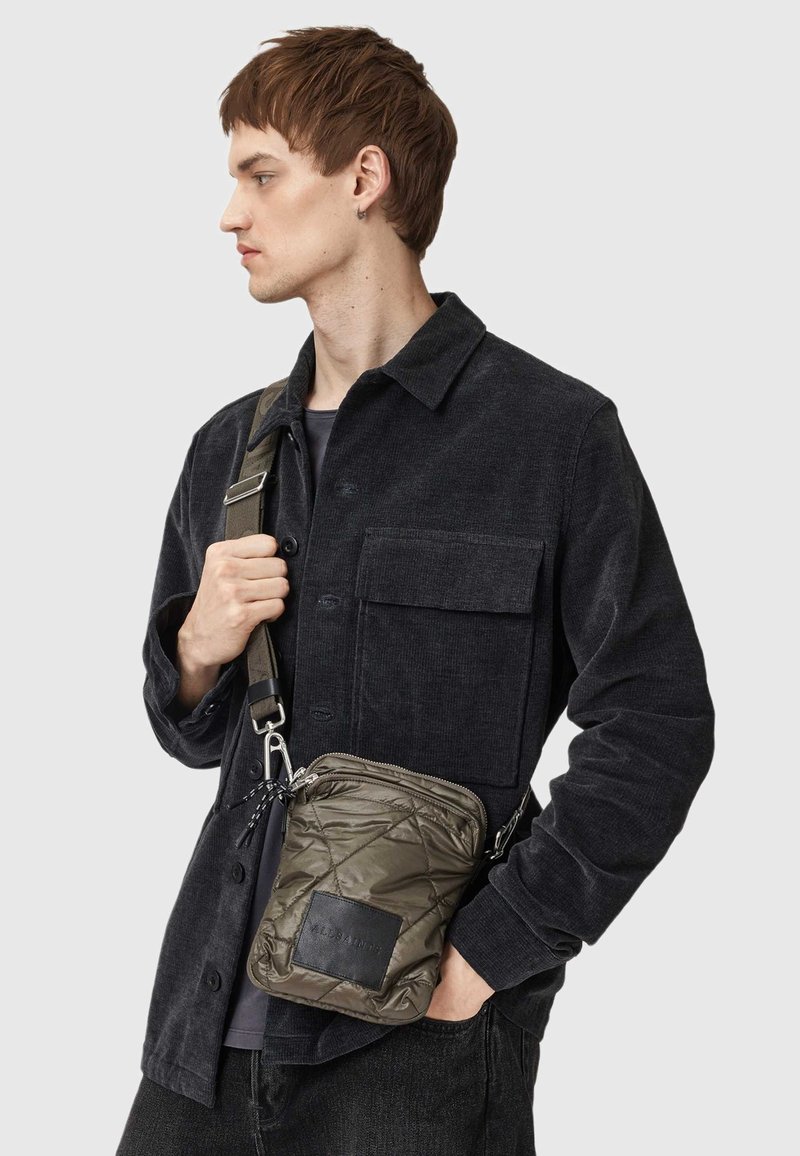 Quilted oliwkowa torba na ramię z czarną skórzaną naszywką, srebrnym zamkiem błyskawicznym i regulowanym paskiem, noszona przez mężczyznę w czarnej kurtce z kordurowej tkaniny.