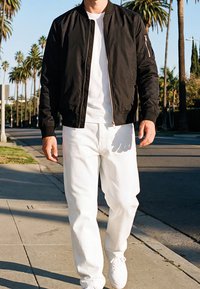 Un homme marche sur le trottoir portant une veste noire, un t-shirt blanc, un pantalon blanc et des baskets blanches, avec des palmiers et un ciel dégagé en arrière-plan.