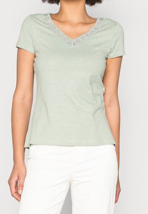 Žena v krátkém světle zeleném topu s výstřihem do V a krajkovým lemem, v kombinaci s bílými kalhotami s vysokým pasem, na jednobarevném pozadí.