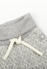 Pantalon de survêtement gris avec une taille douce et texturée, ornée d'un cordon crème, et d'un motif géométrique blanc sur la jambe. Détail du logo présent.