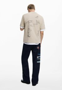 Beige short-sleeve shirt with illustrated designs on the back, paired with dark denim pants featuring patchwork of landscapes and accents.
