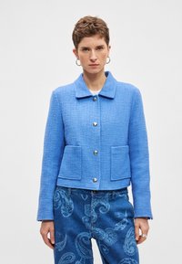 Μπλε υφασμάτινο μπουφάν με γιακά, ασημένια κουμπιά και δύο μπροστινές τσέπες, σε συνδυασμό με μπλε τζιν παντελόνι με σχέδια.