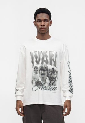 Homme portant une chemise blanche à manches longues avec de grands portraits en noir et blanc de joueurs de football, texte "IVAN Nielsen" à l'avant et texte en cursive sur la manche.