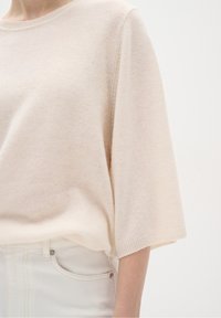 Suéter de punto beige claro con una textura suave, mangas anchas y cuello acanalado. Lleva puesto sobre jeans blancos de cintura alta.