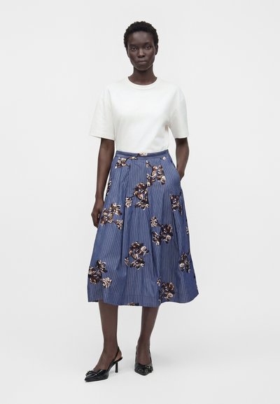 Femme debout portant un haut blanc à manches courtes, une jupe midi bleue à motifs floraux et des chaussures slingback noires à talons bas sur un fond uni.