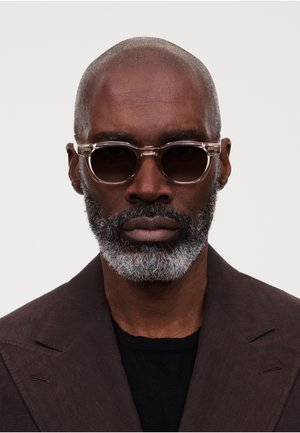 Skallet mann med grått skjegg iført mørke solbriller, svart skjorte og mørk brun blazer mot hvit bakgrunn.