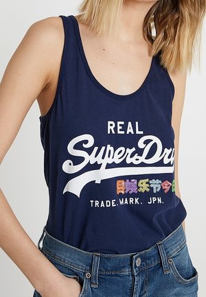 Kobieta w granatowym topie SuperDry z białym i kolorowym japońskim tekstem, włożonym do wysokich, niebieskich dżinsów z wysokim stanem.