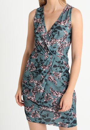 Bezrekawowa turkusowa sukienka w różowy i czarny wzór kwiatowy, z dekoltem w serek, wiązana z przodu, noszona przez stojącą osobę z rękami luźno opuszczonymi.