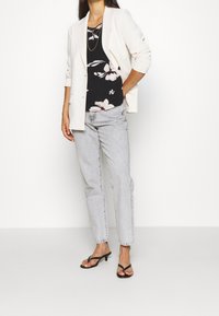 Blazer chiaro sopra una maglietta nera a fiori, abbinato a pantaloni grigi a vita alta e sandali neri con tacco. Collana d'oro semplice in accentuazione.