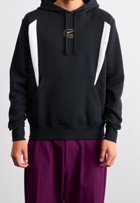 Black hoodie with white panel accents and a gold logo on the chest. Features a front pocket and ribbed cuffs. Smooth fabric texture.