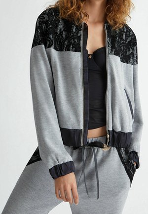 Vrouw draagt een grijze trainingspak met rits en zwarte kantdetails op de schouders, manchetten en broek, gecombineerd met een zwart cropped top eronder.