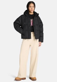 Zwarte donsjas met capuchon, voorzien van een gewatteerd ontwerp, frontknopen en twee zijzakken, gecombineerd met crèmekleurige wijde broek.