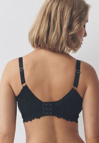 Sort blonde-bh med justerbare stropper, en bølget kant og lukning med hægte og øje. Har detaljerede mønstre og en glat tekstur.