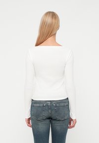 Άτομο με ξανθά μαλλιά που στέκεται με την πλάτη στραμμένη, φορώντας λευκή μπλούζα με μακριά μανίκια και μπλε τζιν παντελόνι με ορατές πίσω τσέπες.