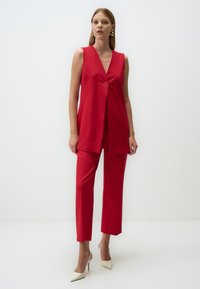 Red sleeveless vest top with a single button closure, paired with matching tailored trousers and beige high-heeled shoes. Smooth fabric texture.