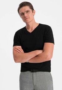 Sort V-hals t-shirt lavet af tekstureret stof, tætsiddende design. Modellen har ternede bukser på, armene krydset, med briller og kort hår.