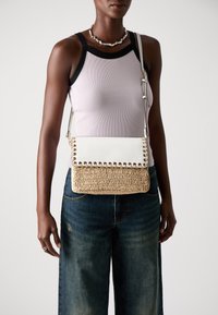 Borsa a tracolla bianca con fondo in paglia intrecciata, dotata di accenti in metallo dorato e finitura testurizzata. Indossata con una maglietta lilla chiara con bordi neri.