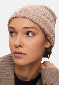 Cappello lavorato a maglia di colore rosa chiaro con texture a coste e logo triangolare decorativo. Il modello indossa un maglione accogliente che mostra uno stile generale.