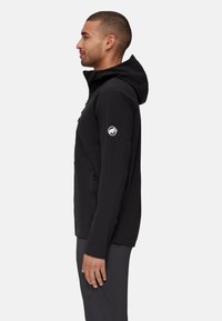Homme portant une veste noire à capuche avec des poches zippées et un logo sur la manche supérieure, se tenant de profil devant un fond uni.