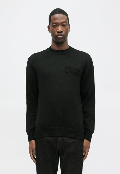 Jaunas vyras vilki juodą, raukšlėtą apvalią kaklo megztinį su iškilniu logotipu ant krūtinės, stovėdamas su atsipalaidavusiais rankomis prieš paprastą baltą foną.