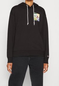 Felpa con cappuccio nera realizzata in cotone, con una patch grafica multicolore sul petto. Rifinita con lacci bianchi e polsini a coste.
